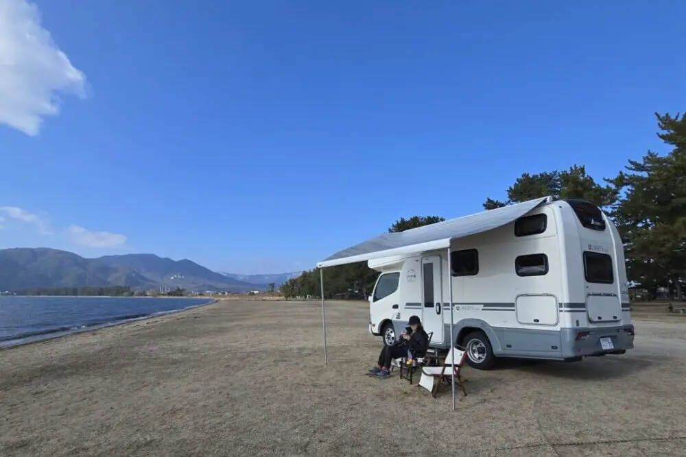 砂浜でダイネットを広げた車の前でイスに座っている女性